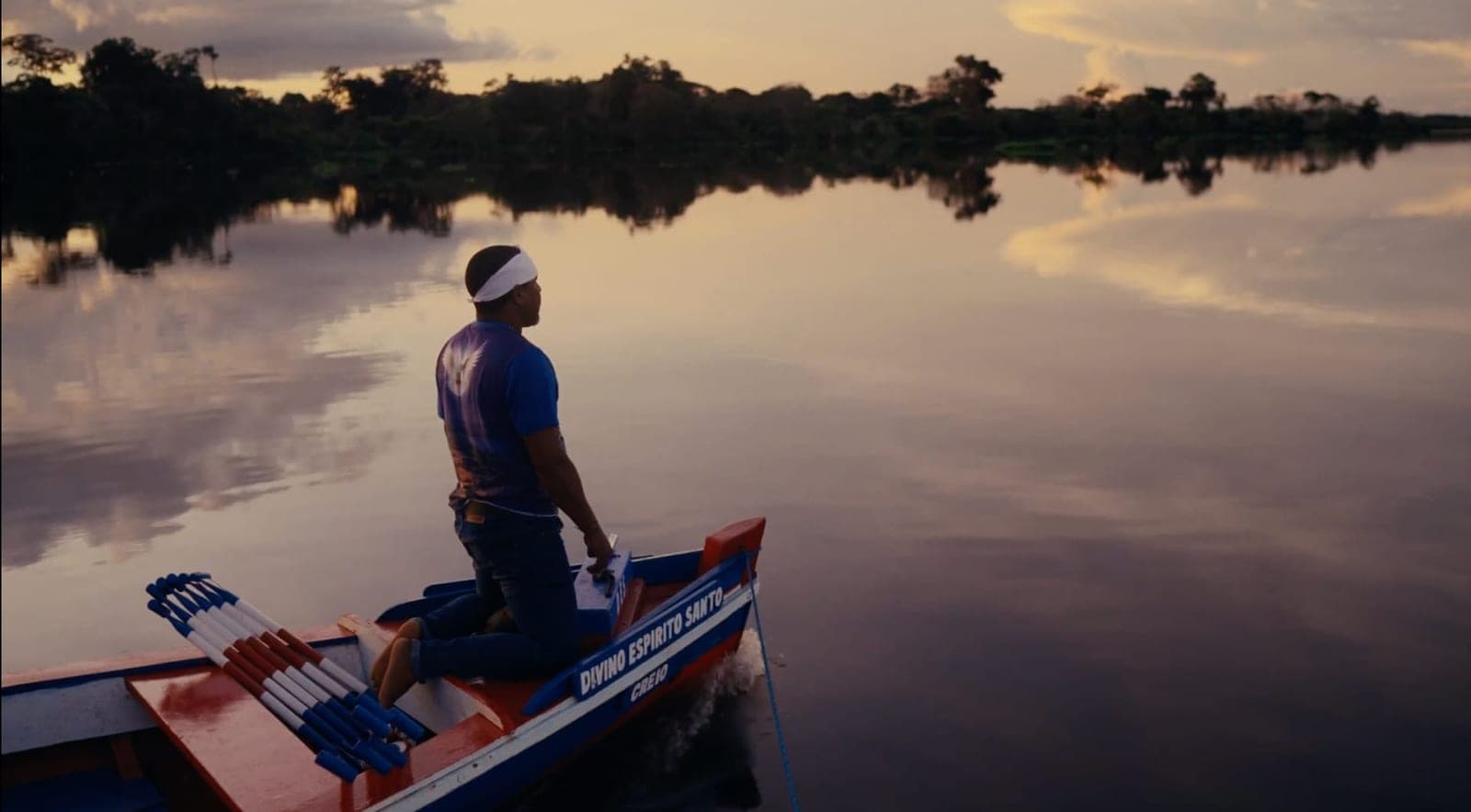 Homem de pé na proa de um barco, no rio, ao entardecer, com a mata refletida na água.
