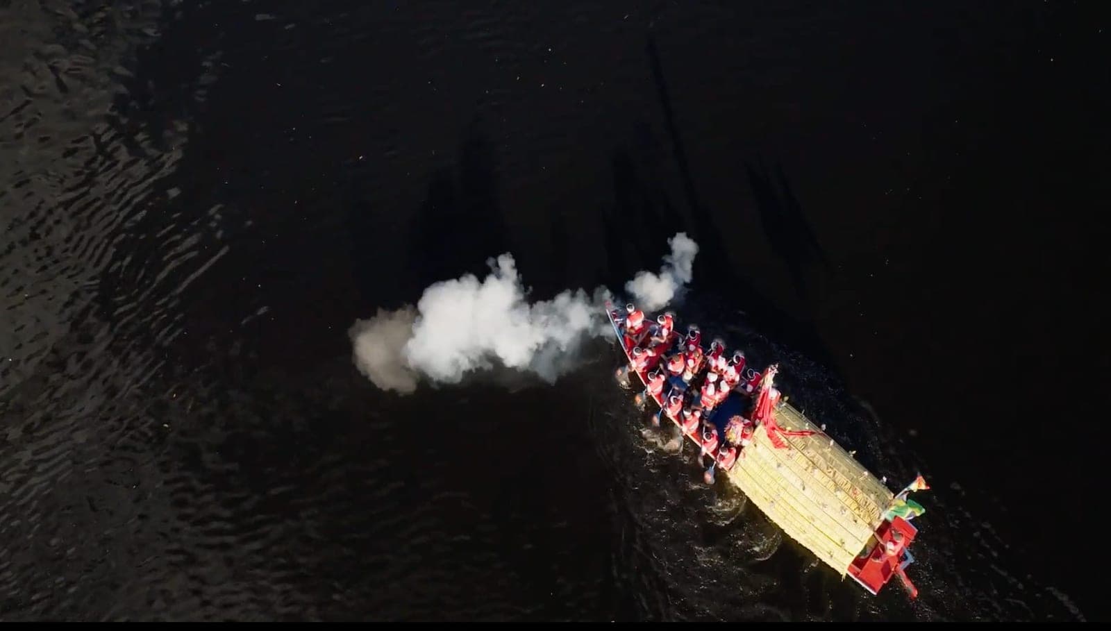 Vista aérea, à noite, de um barco enfeitado no rio, com a fumaça de fogos saindo dele.