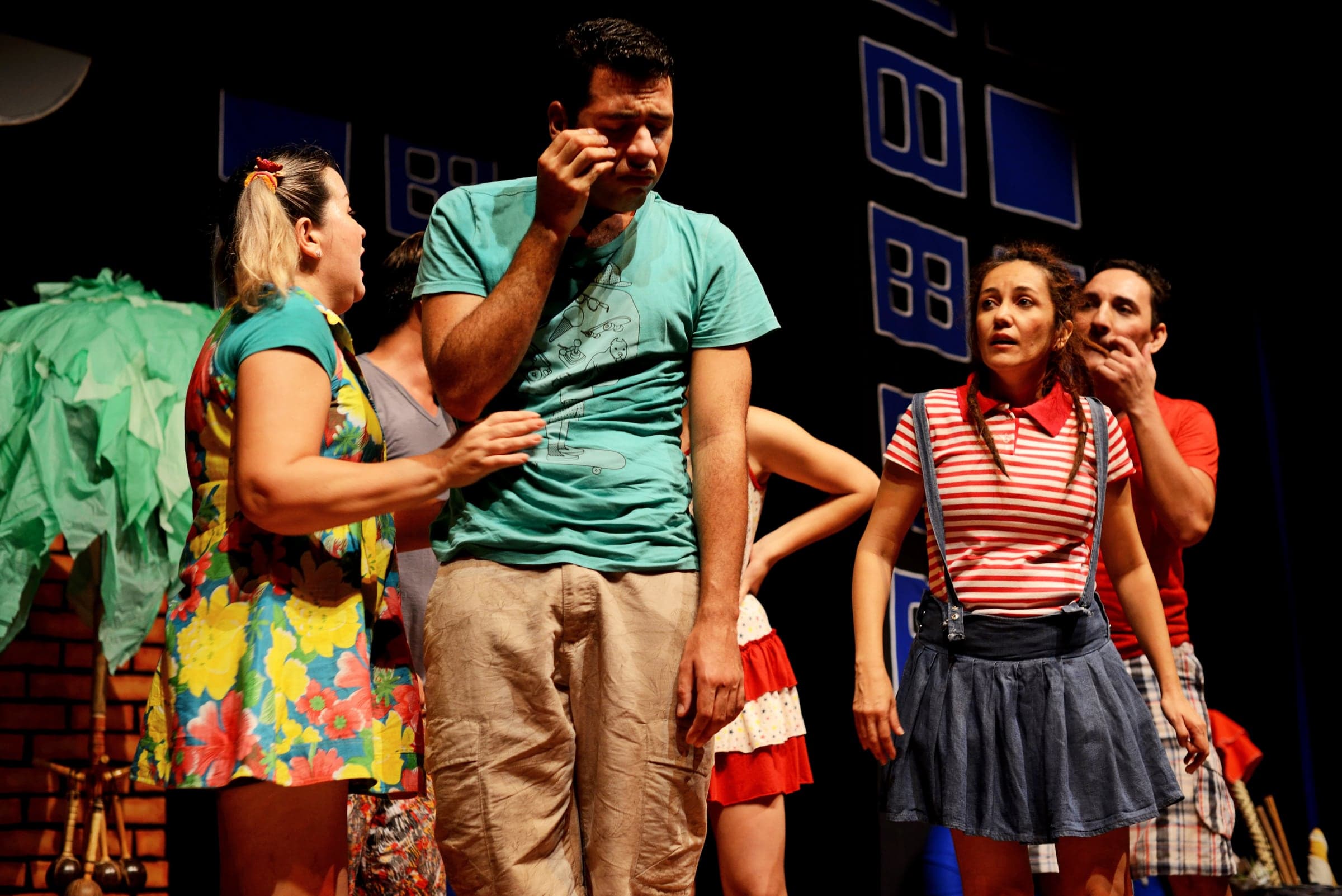 Cena de comédia com o elenco em roupas coloridas no palco.