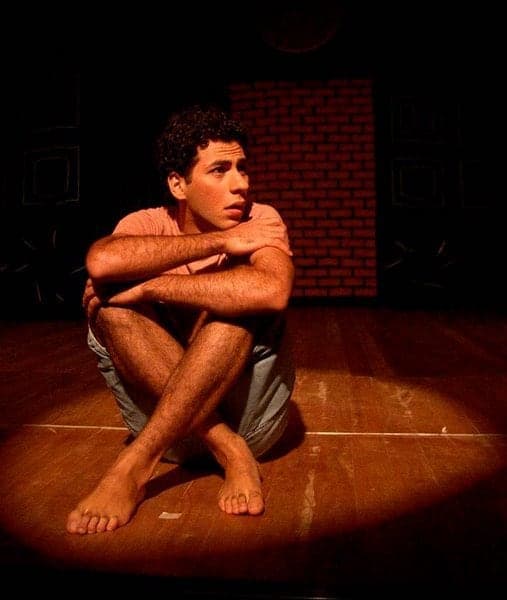 Sentado no chão do palco, abraçando os joelhos, sob um foco de luz.