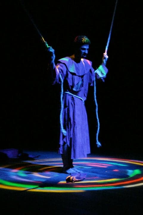 O Frei de pé, de túnica azul, segurando duas cordas sobre um círculo de luz colorida.