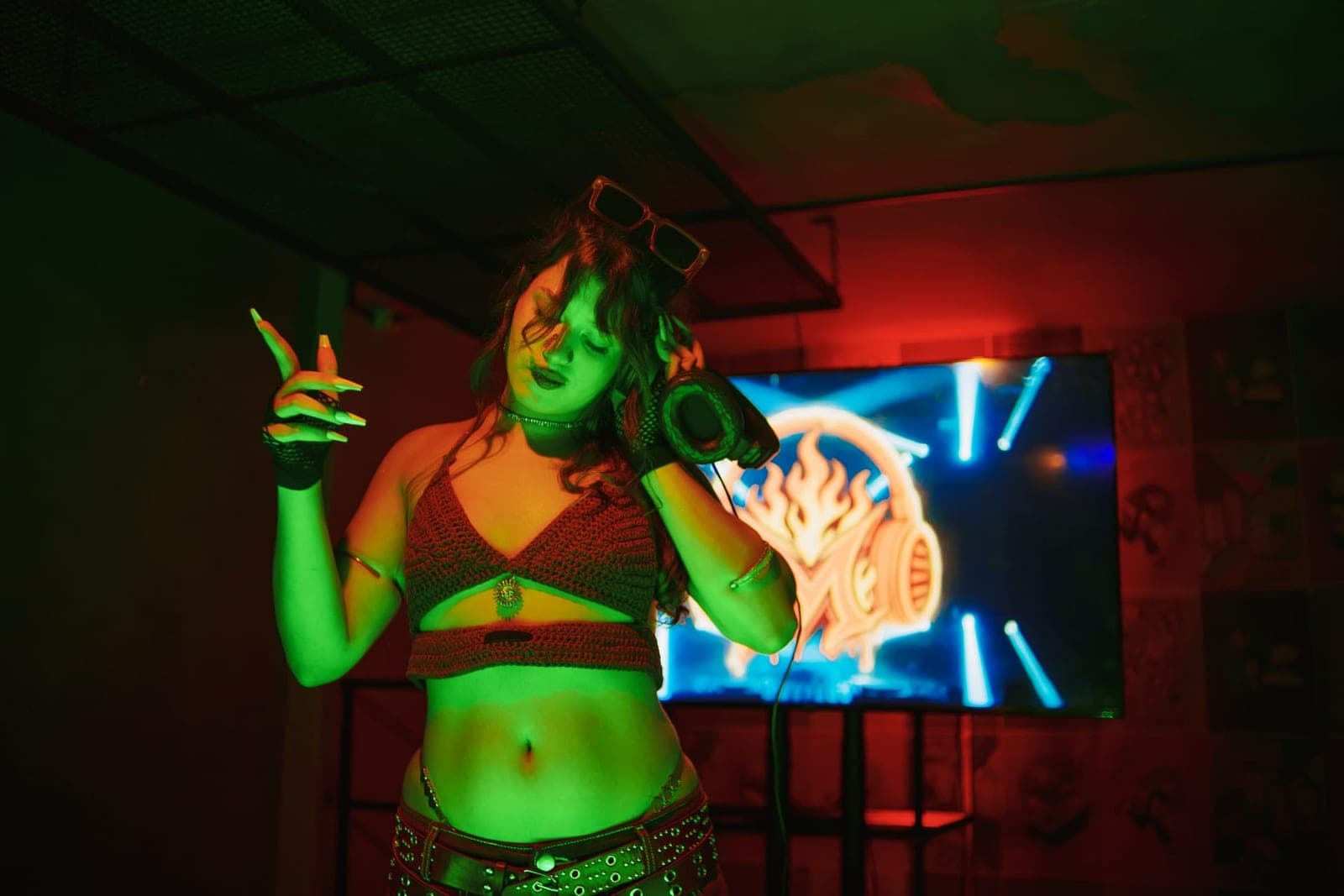 Milena dança sob luz verde, com fones de ouvido no pescoço, diante de uma tela iluminada.