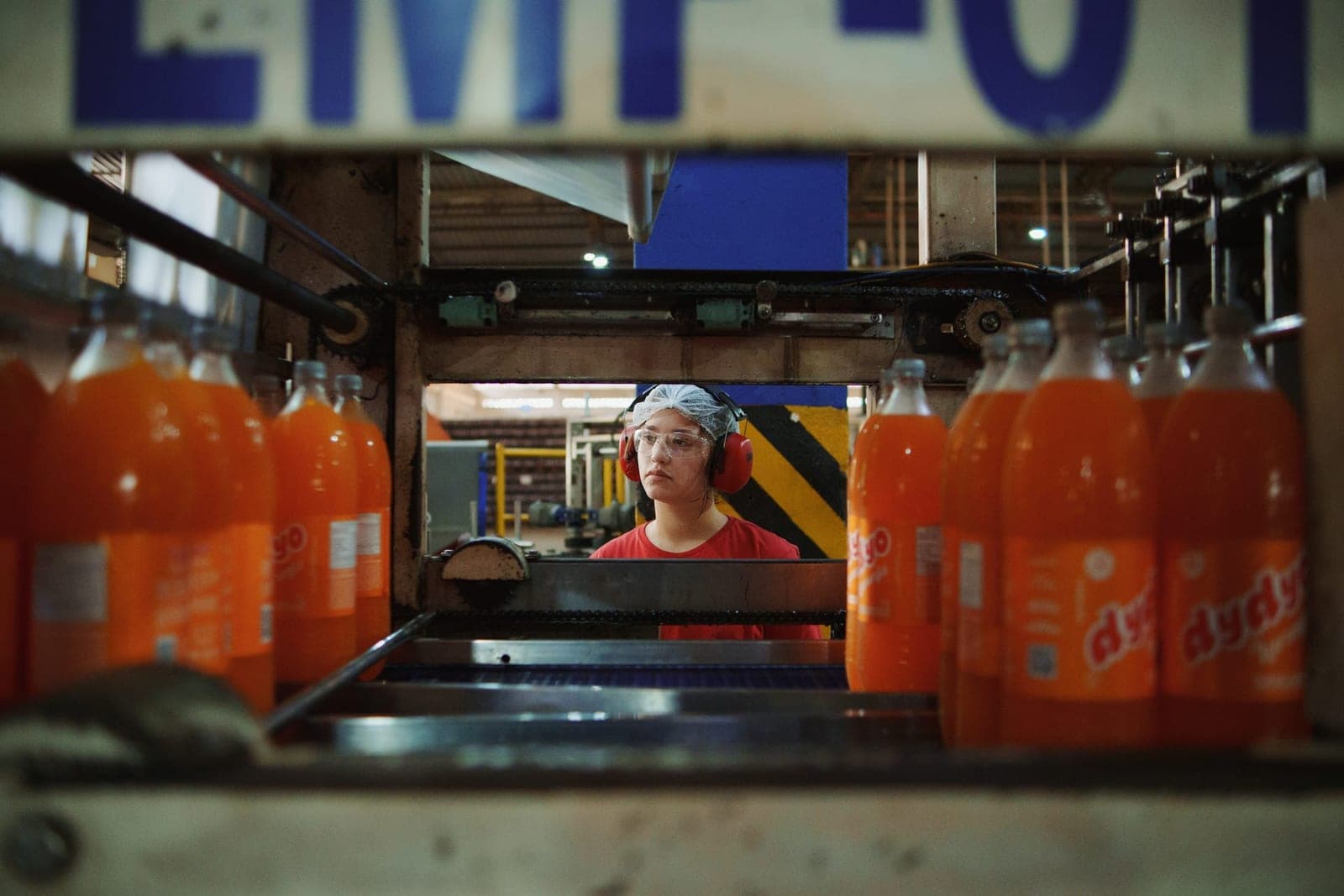 Milena, de protetor auricular, atrás da linha de garrafas de refrigerante laranja de uma fábrica.