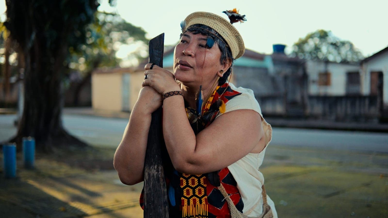 Mulher indígena de cocar e colares segurando um objeto junto ao rosto, numa rua.