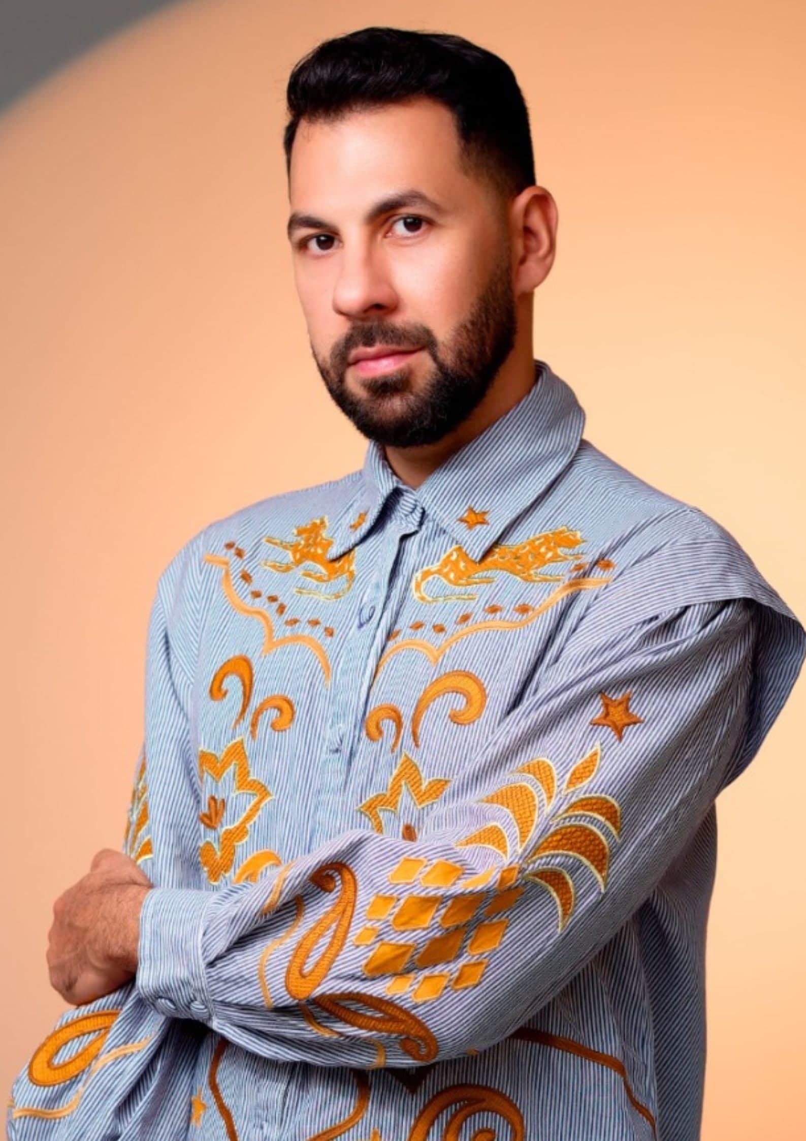 Retrato de Juraci Júnior em camisa listrada com bordados laranja, diante de um fundo de luz quente.