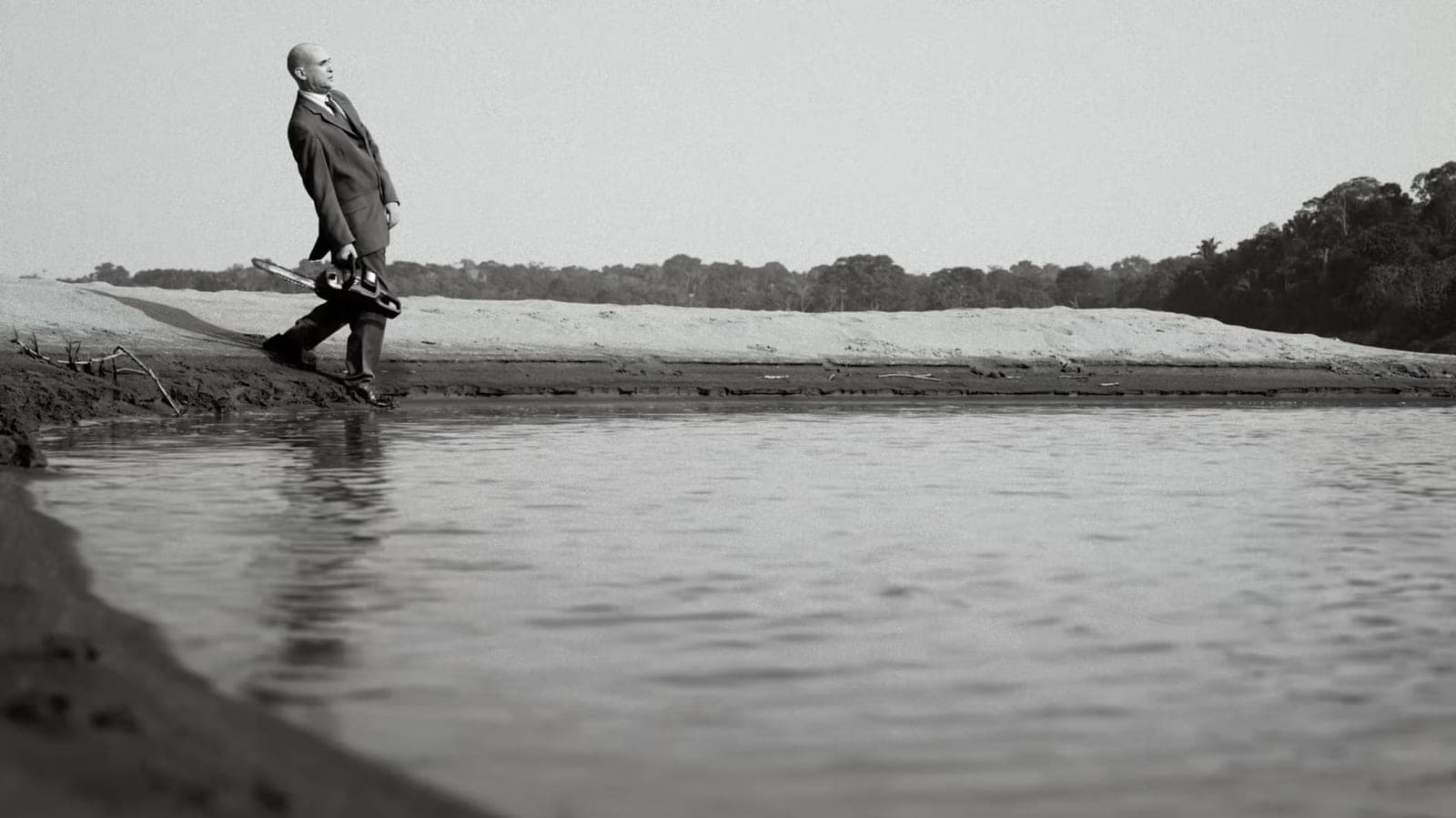 Em preto e branco, homem de terno carregando uma motosserra pela margem de areia do rio.