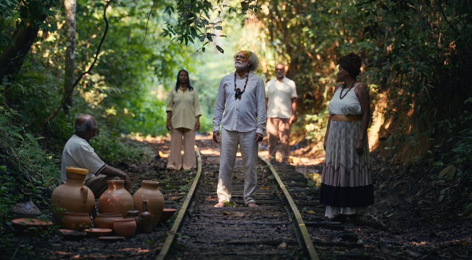 Pessoas paradas sobre um trilho de trem no meio da mata, com potes de barro no chão.