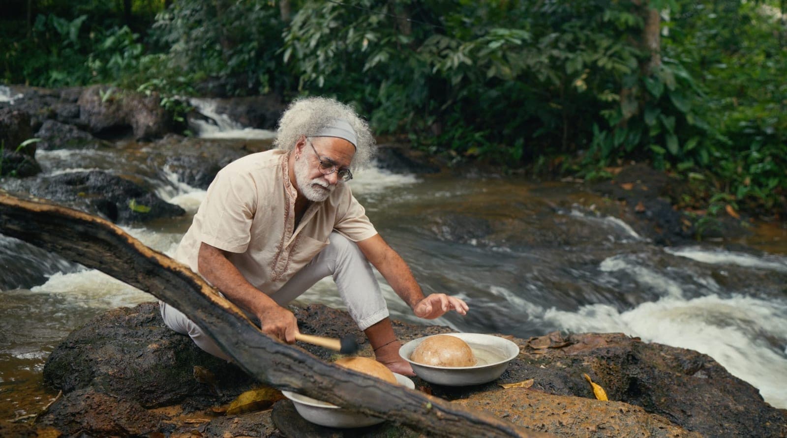 Homem de barba branca agachado numa pedra junto a uma cachoeira, com cuias de barro.