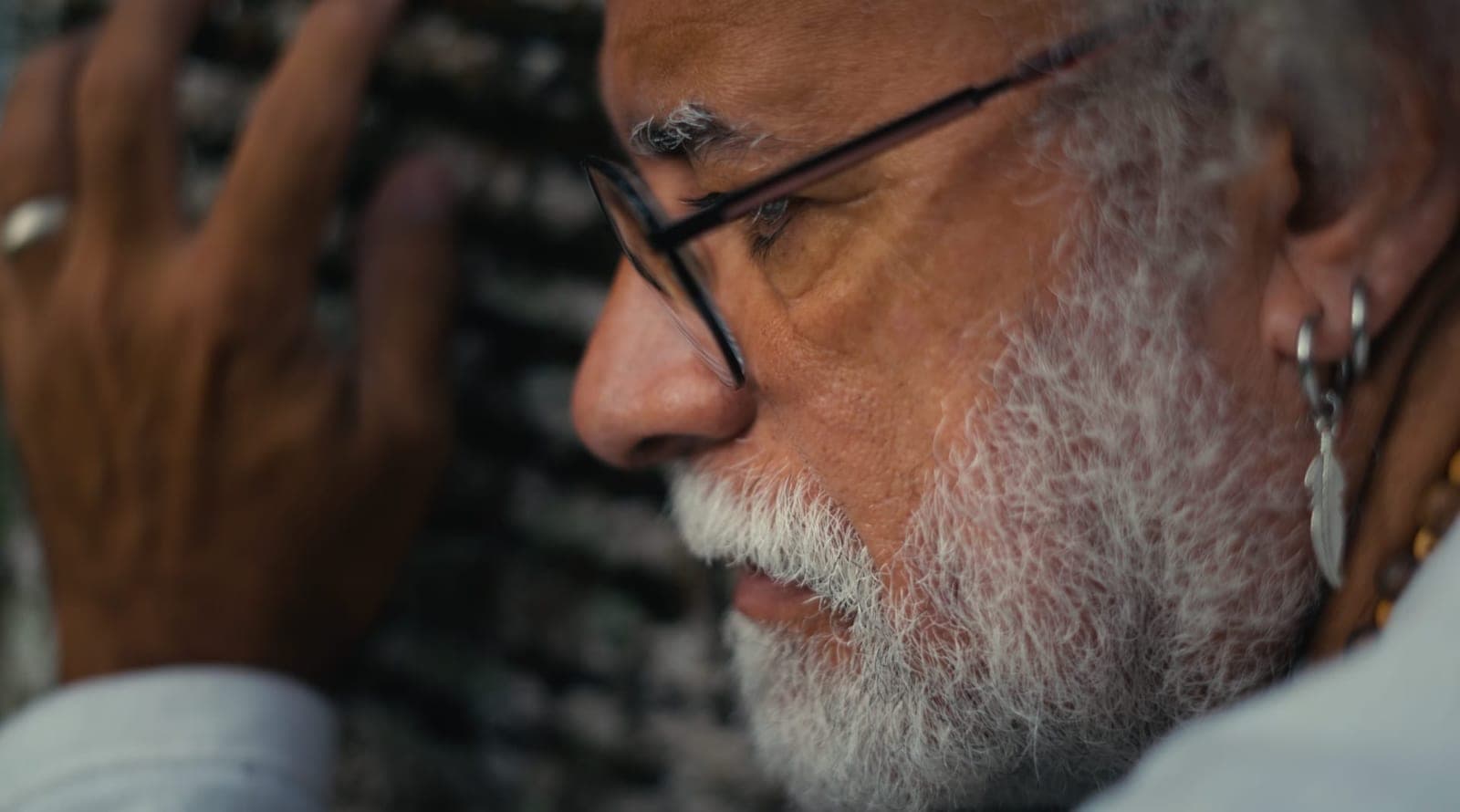 Perfil de um homem de óculos e barba branca, com brinco, encostado numa parede.